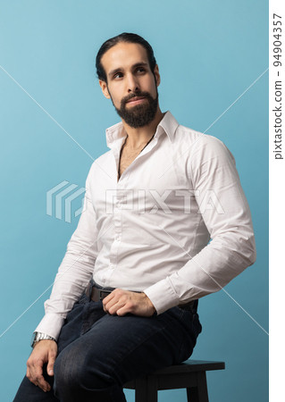 Unsmiling serious dark haired attractive man with beard wearing white shirt and black trousers, looking away with attentive look while sitting on chair. Indoor studio shot isolated on blue background. 94904357