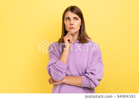 Portrait of young adult thoughtful woman standing holding chin, looking away, thinks about something important, wearing purple hoodie. Indoor studio shot isolated on yellow background. Portrait of young adult thoughtful woman standing holding chin, looking away, thinks about something important, wearing purple hoodie. Indoor studio shot isolated on yellow background. 94904381