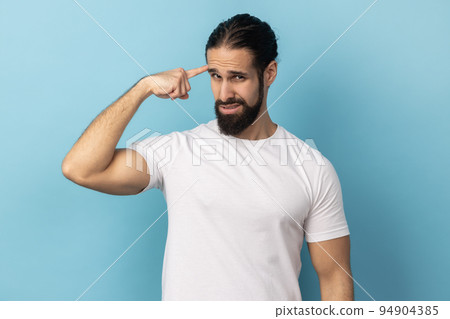 This is insane idea. Portrait of man with beard showing stupid gesture with finger at her temple, displeased with reckless silly joke, out of mind. Indoor studio shot isolated on blue background. 94904385