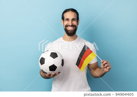 Portrait of smiling man with beard wearing white T-shirt supporting german soccer team on championship, cheering and greeting, patriotism. Indoor studio shot isolated on blue background. 94904405