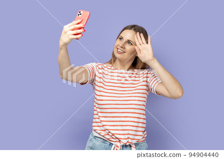 Portrait of cheerful blond woman wearing striped T-shirt with mobile phone phone and having video call, having hi or bye with hand. Indoor studio shot isolated on purple background. 94904440