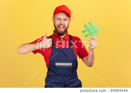 Portrait of excited handyman wearing blue overalls standing holding green hashtag and showing thumb up, looking at camera with smile. Indoor studio shot isolated on yellow background. 94904446