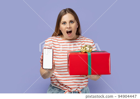 Amazed excited woman wearing striped T-shirt holding red present box and showing cell phone with white screen with copy space for advertisement. Indoor studio shot isolated on purple background. 94904447