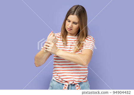 Unhappy sick blond woman wearing striped T-shirt touching painful hand, suffering trauma, sprain wrist, feeling ache of carpal tunnel syndrome. Indoor studio shot isolated on purple background. Unhappy sick blond woman wearing striped T-shirt touching painful hand, suffering trauma, sprain wrist, feeling ache of carpal tunnel syndrome. Indoor studio shot isolated on purple background. 94904482