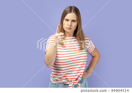 Portrait of blond woman wearing striped T-shirt pointing finger at camera and looking with dissatisfied suspicious expression, warning about troubles. Indoor studio shot isolated on purple background. Portrait of blond woman wearing striped T-shirt pointing finger at camera and looking with dissatisfied suspicious expression, warning about troubles. Indoor studio shot isolated on purple background. 94904514