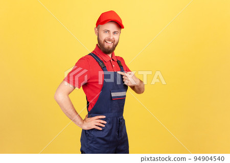 Side view of proud successful worker man standing and pointing at himself, bragging about the result of his work, wearing overalls and red cap. Indoor studio shot isolated on yellow background. Side view of proud successful worker man standing and pointing at himself, bragging about the result of his work, wearing overalls and red cap. Indoor studio shot isolated on yellow background. 94904540