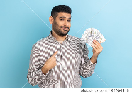 Portrait of young adult businessman with beard holding dollar bills and pointing at himself with pride, looking at camera, wearing striped shirt. Indoor studio shot isolated on blue background. Portrait of young adult businessman with beard holding dollar bills and pointing at himself with pride, looking at camera, wearing striped shirt. Indoor studio shot isolated on blue background. 94904546