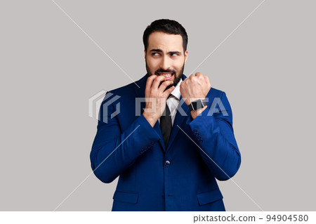 Nervous bearded man biting finger nails and looking at camera, showing his wrist clock, worried about deadline, wearing official style suit. Indoor studio shot isolated on gray background. Nervous bearded man biting finger nails and looking at camera, showing his wrist clock, worried about deadline, wearing official style suit. Indoor studio shot isolated on gray background. 94904580