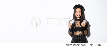 Image of beautiful asian woman in gothic lace dress and black wreath, looking aside pleased and smiling, holding mobile phone with credit card Image of beautiful asian woman in gothic lace dress and black wreath, looking aside pleased and smiling, holding mobile phone with credit card 94904615