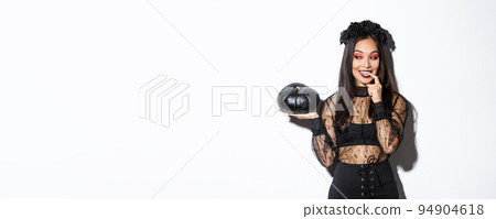 Image of sexy asian woman in witch costume looking at black pumpking and smiling, standing over white background 94904618