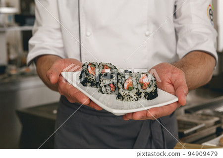 Close up of chef of japanies restaurant showing plate with sushi standing on kitchen Close up of chef of japanies restaurant showing plate with sushi standing on kitchen 94909479