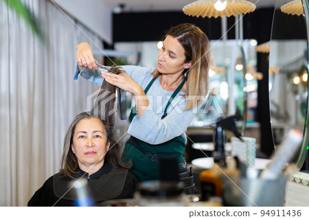 Focused woman hairdresser cutting hair of elderly female client Focused woman hairdresser cutting hair of elderly female client 94911436
