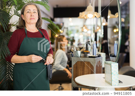 Confident elderly female hairdresser standing in hair salon Confident elderly female hairdresser standing in hair salon 94911805