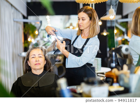 Focused woman hairdresser cutting hair of elderly female client 94911817