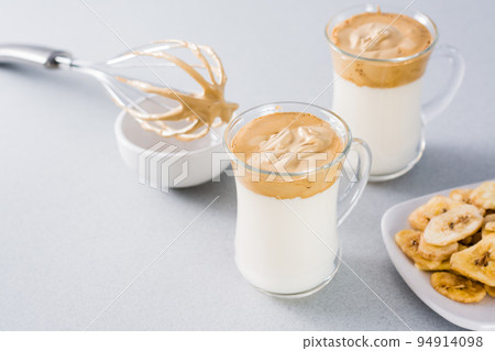 Quarantine trendy cuisine. Two cups with dalgona coffee and banana chips and a whisk on a gray background. 94914098