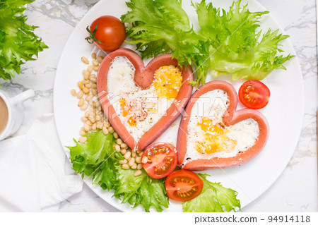 Romantic breakfast. Fried eggs in heart shaped sausages, lettuce and cherry tomatoes on a plate on the table. Top view. Close-up Romantic breakfast. Fried eggs in heart shaped sausages, lettuce and cherry tomatoes on a plate on the table. Top view. Close-up 94914118