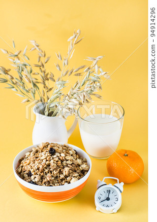 Healthy hearty breakfast. Baked granola in bowl, glass of milk, orange and alarm clock on yellow background. Vertical view 94914296