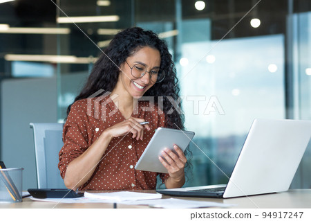Young beautiful Latin American woman working inside modern office building, paperwork woman uses tablet computer and laptop, accountant calculates reports and accounts, financial documents. Young beautiful Latin American woman working inside modern office building, paperwork woman uses tablet computer and laptop, accountant calculates reports and accounts, financial documents. 94917247