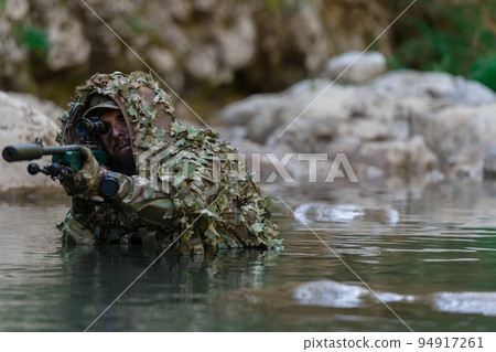 A military man or airsoft player in a camouflage suit sneaking the river and aims from a sniper rifle to the side or to target. 94917261
