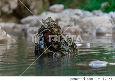 A military man or airsoft player in a camouflage suit sneaking the river and aims from a sniper rifle to the side or to target. A military man or airsoft player in a camouflage suit sneaking the river and aims from a sniper rifle to the side or to target. 94917263