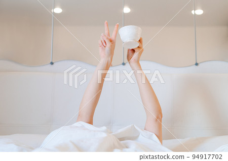 Young woman with a coffee mug in bed with white linens. Minimal happy morning Young woman with a coffee mug in bed with white linens. Minimal happy morning 94917702