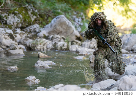 A military man or airsoft player in a camouflage suit sneaking the river and aims from a sniper rifle to the side or to target. 94918036