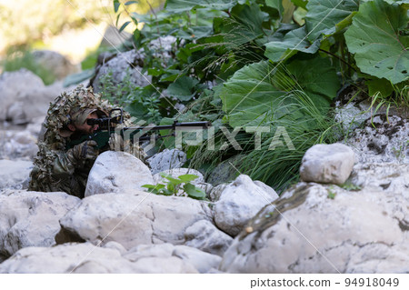A military man or airsoft player in a camouflage suit sneaking the river and aims from a sniper rifle to the side or to target. 94918049