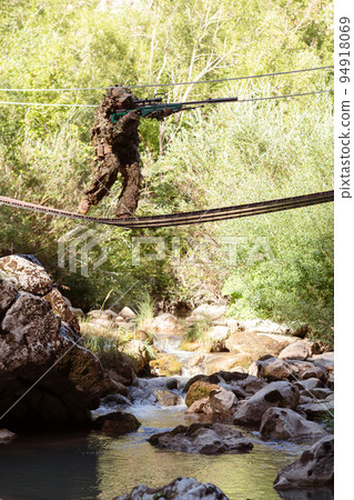 A military man or airsoft player in a camouflage suit sneaking the rope bridge and aims from a sniper rifle to the side or to target. A military man or airsoft player in a camouflage suit sneaking the rope bridge and aims from a sniper rifle to the side or to target. 94918069
