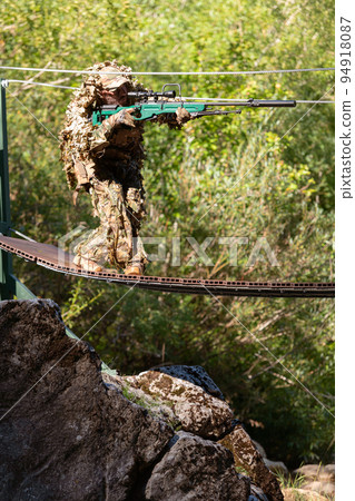 A military man or airsoft player in a camouflage suit sneaking the rope bridge and aims from a sniper rifle to the side or to target. A military man or airsoft player in a camouflage suit sneaking the rope bridge and aims from a sniper rifle to the side or to target. 94918087