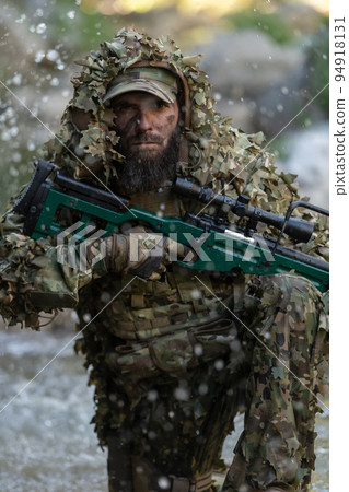 A military man or airsoft player in a camouflage suit sneaking the river and aims from a sniper rifle to the side or to target. 94918131