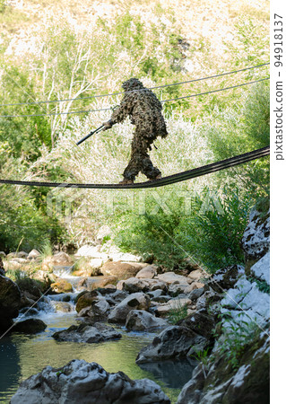 A military man or airsoft player in a camouflage suit sneaking the rope bridge and aims from a sniper rifle to the side or to target.  94918137