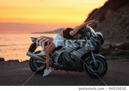 Freedom and travel. Caucasian young woman sexy sitting on motorcycle. Golden epic sunset and motorbike on the background. Concept of World Motorcyclist Day and moto travel Freedom and travel. Caucasian young woman sexy sitting on motorcycle. Golden epic sunset and motorbike on the background. Concept of World Motorcyclist Day and moto travel 94919830