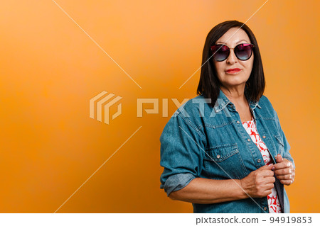 Women and advertising. Mature brunette woman in glasses and denim shirt posing on orange background. Horisontal orientation. Copy space Women and advertising. Mature brunette woman in glasses and denim shirt posing on orange background. Horisontal orientation. Copy space 94919853