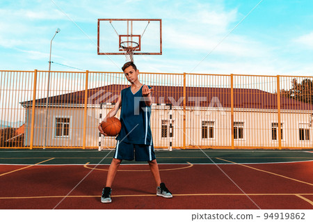 Sports and basketball. A young teenager in a blue tracksuit confidently poses with a ball, standing on the Playground, and beckons. Blue sky and building in the background Sports and basketball. A young teenager in a blue tracksuit confidently poses with a ball, standing on the Playground, and beckons. Blue sky and building in the background 94919862