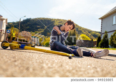 njury. A teenage boy lies on the ground next to a scooter, rubbing his bruised shoulder. Outdoor. Concept of extreme sports and youth activity njury. A teenage boy lies on the ground next to a scooter, rubbing his bruised shoulder. Outdoor. Concept of extreme sports and youth activity 94919865