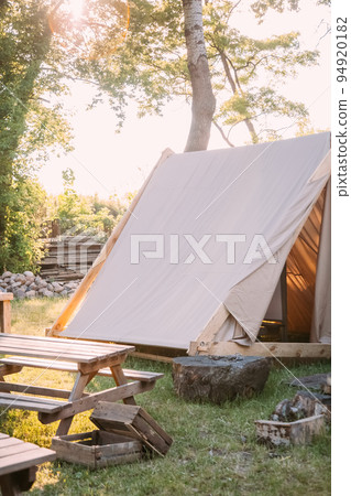Rural camping. Rest zone. Close nature. Canvas tent with picnic desk on sunshine yard and wooden boxes outdoor. Rural camping. Rest zone. Close nature. Canvas tent with picnic desk on sunshine yard and wooden boxes outdoor. 94920182