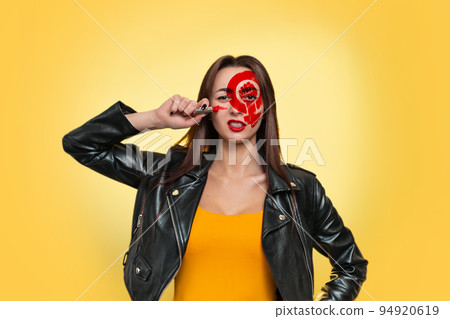 Feminism and the concept of equal rights and independence for women. A Caucasian young woman in a leather jacket draws war paint on her face with lipstick. Yellow background. Copy space Feminism and the concept of equal rights and independence for women. A Caucasian young woman in a leather jacket draws war paint on her face with lipstick. Yellow background. Copy space 94920619