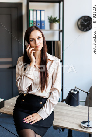 Business talk. Cheerful young beautiful woman talking on mobile phone and looking to the window while leaning on the table at her working place 94921233