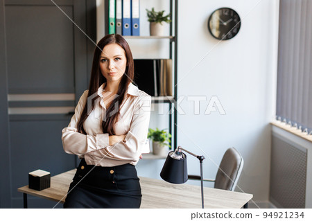 Pleasant young female assistant, manager standing on workplace in office near window, with pleased confident expression, cross hands over chest showing readiness, business and women concept 94921234