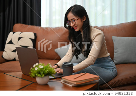 Beautiful Asian female in eyeglasses using laptop while sitting on the couch in the living room. 94921338
