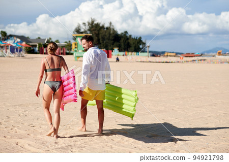 Rear view of young couple with swimming mattresses walking along the beach Rear view of young couple with swimming mattresses walking along the beach 94921798