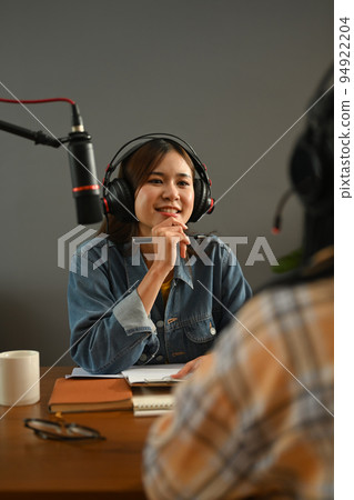 Friendly radio host in headphone discussing various topics with her guest while streaming live audio podcast from home studio Friendly radio host in headphone discussing various topics with her guest while streaming live audio podcast from home studio 94922204
