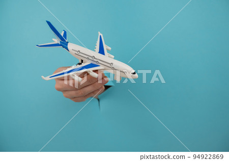 A female hand sticking out of a hole from a blue background holds a model of an airplane. A female hand sticking out of a hole from a blue background holds a model of an airplane. 94922869