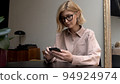 Adult woman in eyeglasses, pink shirt holding smartphone in hands, writing a message smiling and 94924974