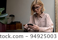 Adult woman in eyeglasses, pink shirt holding smartphone in hands, writing a message smiling and 94925003