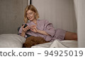 Caucasian blonde woman smiling and typing in her device early in the morning, pet lying on bed with 94925019