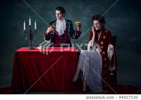 Portrait of man and woman in image of medieval vampires over dark green background. Man eating burger, woman talking on phone Portrait of man and woman in image of medieval vampires over dark green background. Man eating burger, woman talking on phone 94927330