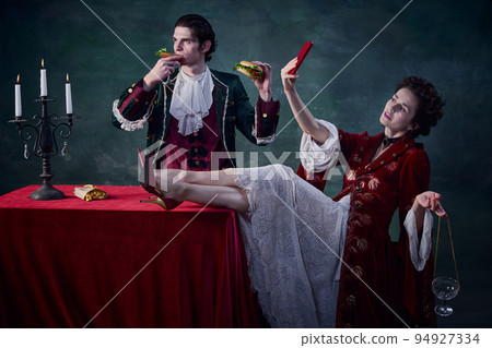 Portrait of man and woman in image of medieval vampires over dark green background. Man eating burger, woman drinking and taking selfie 94927334