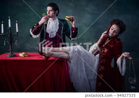 Portrait of man and woman in image of medieval vampires over dark green background. Man eating burger and drinking bloody mary, woman talking on phone 94927335