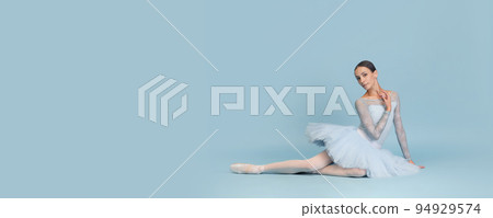 Portrait of tender young ballerina dancing, performing, sitting on floor isolated over blue studio background. Flyer image 94929574